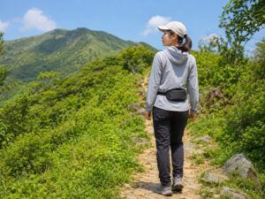 女性ハイカーがウエストポーチを後ろに装着し、背面を空けて夏の登山道を歩く様子