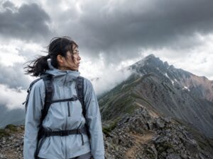 強風と雲が迫る高山稜線でレインウェアを着る女性登山者